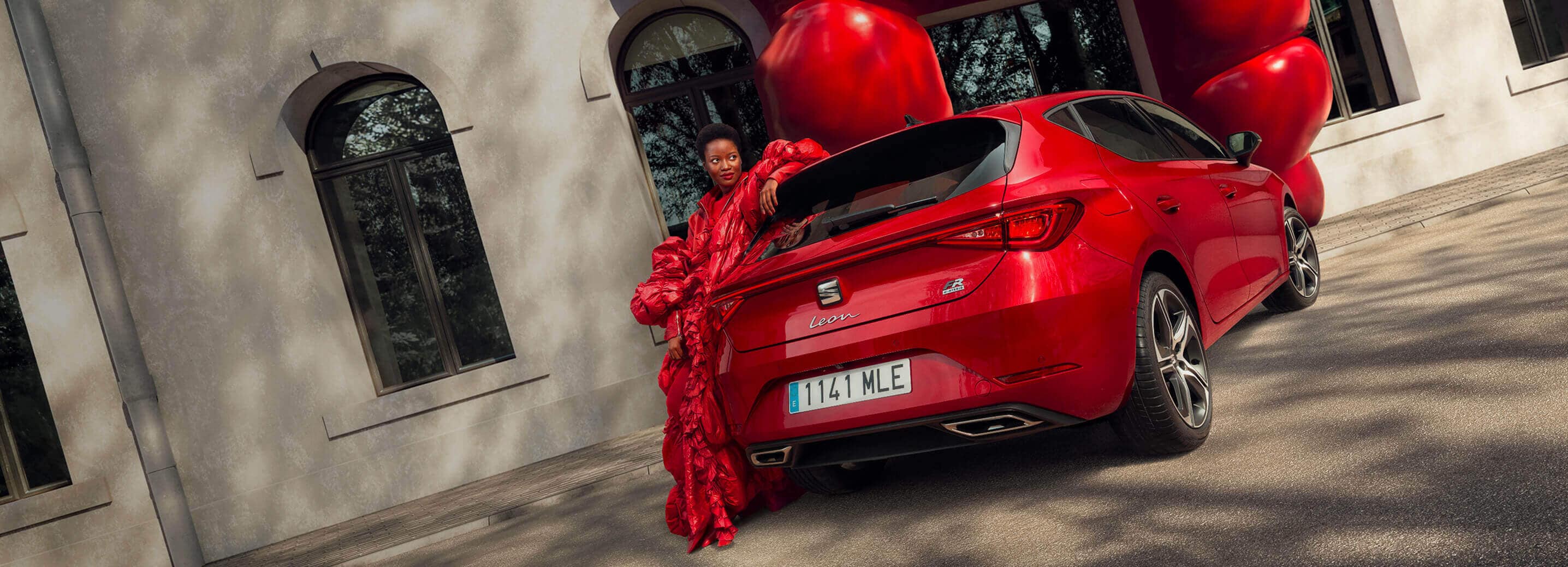 woman standing next to a red seat leon by a modern brick wall, ready for a test drive that combines style and performance